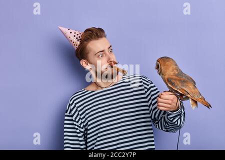 Aufgeregt positive lustige Mann in Hut und mit Party-Horn im Mund Blick auf den Vogel. Unterhaltung. Freude, Gegenwart. Isoliert blauen Hintergrund, Studio Stockfoto