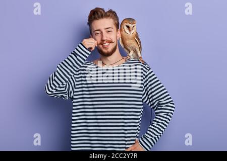 Lächelnder hübscher Mann mit einer Eule auf seiner Schulter sitzend, seinen Schnurrbart berührend. Isolierter blauer Hintergrund Studioaufnahme. Stockfoto