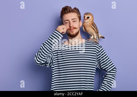 Fröhlicher lustiger schlauer Kerl im gestreiften Pullover, der Geld vom Touristen nimmt, weil er ein Pictyre mit einer Eule nimmt, isolierter blauer Hintergrund, Studioaufnahme, Busin Stockfoto