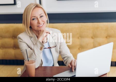 Fröhliche blonde Frau, die mit Laptop im Café sitzt. Nahaufnahme Foto. Lifestyle, Job, Beauty-Konzepte Stockfoto