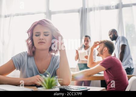 Selektiver Fokus der enttäuschten Geschäftsfrau sitzt am Tisch, während multikulturell Kollegen, die im Büro arbeiten Stockfoto