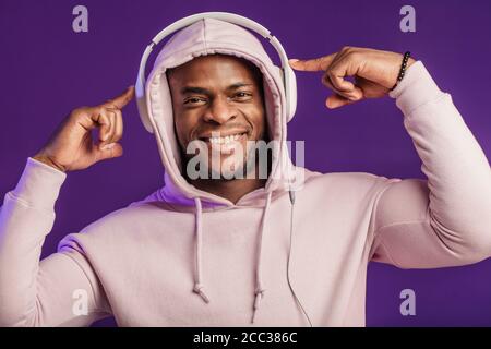 Junge afrikanische Mann in Hoodie, in der Sinn für Mode eines street Urchin, drehte sich ein wenig in der Kopfhörer auf der violette Hintergrund isoliert, holding Ohrhörer Stockfoto