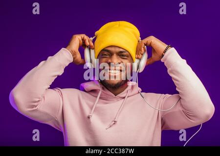 Junge afrikanische Mann in Hoodie, in der Sinn für Mode eines street Urchin, drehte sich ein wenig in der Kopfhörer auf der violette Hintergrund isoliert, holding Ohrhörer Stockfoto