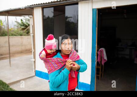 Porträt einer Mutter, die ein Baby auf dem Rücken trägt, in ihrem Zuhause in Manchay Alto, Bezirk Pachamac, Peru. Stockfoto