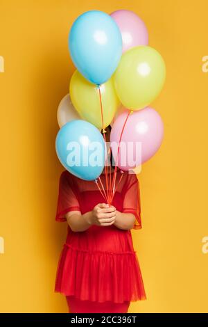 Schwangere Frau im roten Outfit verdeckt Gesicht mit bunten festlichen Ballons auf gelb Stockfoto
