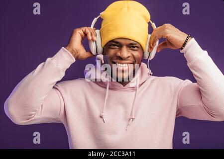 Junge afrikanische Mann in Hoodie, in der Sinn für Mode eines street Urchin, drehte sich ein wenig in der Kopfhörer auf der violette Hintergrund isoliert, holding Ohrhörer Stockfoto