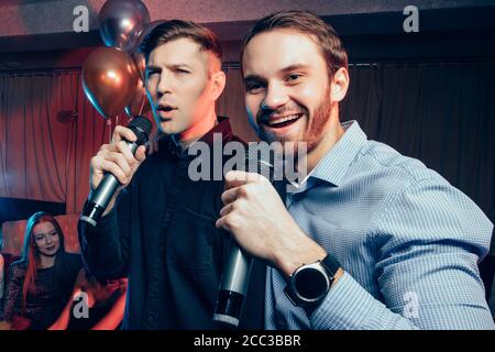 Zwei junge kaukasische Männer in Hemden singen in Mikrofon in Karaoke-Bar, Spaß haben, feiern. Urlaub, Freizeit, Party-Konzept Stockfoto