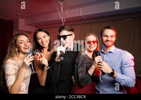 Junge kaukasische Freunde haben Spaß zusammen in Karaoke-Bar, tragen Kleid und T-Shirt, trendige moderne Kleidung. Drinnen von Karaoke-Raum mit Neonlicht Stockfoto