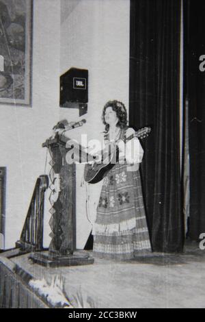 Feine Vintage-Schwarz-Weiß-Fotografie aus den 1970er Jahren von einer Frau, die auf einem Podium eine Gitarre spielt und ihr traditionelles osteuropäisches Gewand trägt. Stockfoto