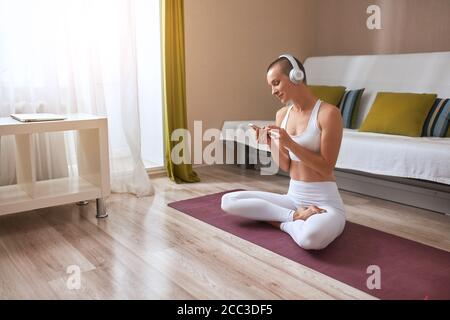 Yogi Attraktive Mädchen arbeiten zu Hause in hellen Raum. Sitzend in der Variante der leichten Haltung auf der speziellen Matte, Sukhasana. Smartphone in Händen und Kopf Stockfoto