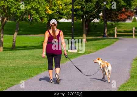 Isoliertes Nahaufnahme-Foto einer jungen blonden Frau in Sportkleidung, während sie mit ihrem Hund im Park läuft. Der hellbraune weiße Hund ist an der Leine. Die w Stockfoto