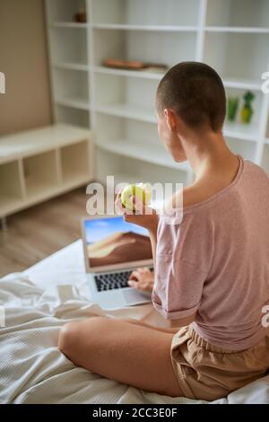 Kurzhaarige Frau im Schlafanzug sitzen mit Laptop und essen grünen Apfel in hellen Raum zu Hause. Rückansicht Stockfoto