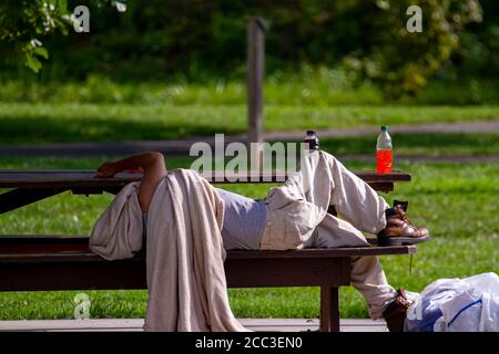 Ein obdachloser Kaukasusmann schläft auf einem Picknicktisch in einem schattigen Bereich eines Parks. Er bedeckt seinen Kopf mit einer schmutzigen Decke. Seine Habseligkeiten sind in Stockfoto