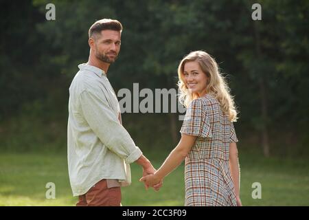Waist-up-Porträt von romantischen erwachsenen Paar Hände halten und Blick auf Kamera über Schulter beim Gehen auf grünem Gras in der Natur Landschaft, Kopie SPAC Stockfoto
