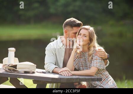 Portrait von romantischen erwachsenen Paar umarmen, während sitzen am Tisch im Freien am See, konzentrieren sich auf lächelnde blonde Frau Blick auf Kamera, kopieren Raum Stockfoto