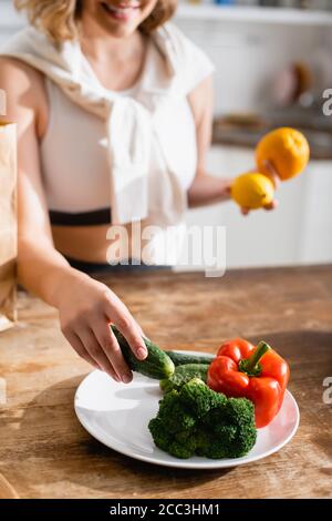 Beschnittene Ansicht der Frau, die Gurke in der Nähe von Teller mit Brokkoli hält Und Paprika Stockfoto