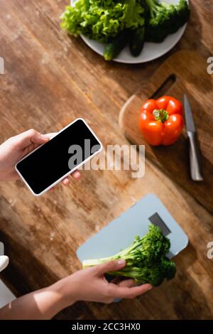 Draufsicht der Frau, die Brokkoli in der Nähe von Küchenwaagen hält und Smartphone mit leerem Bildschirm Stockfoto