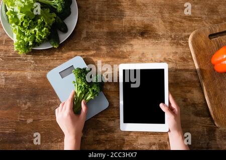 Draufsicht der Frau, die Brokkoli in der Nähe von Küchenwaagen hält und Digitales Tablet mit leerem Bildschirm Stockfoto