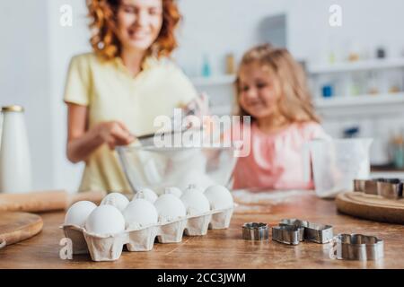 Selektiver Fokus der Frau, die Mehl in eine Glasschüssel neben der Tochter sieben, Hühnereier im Tablett und Ausstechformen auf dem Tisch Stockfoto