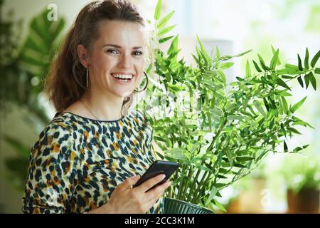 Porträt der glücklichen 40 Jahre alte Frau in Bluse mit grünen Zimmerpflanze mit Smartphone-App und Dekoration zu Hause in modernen Zuhause an sonnigen Tagen. Stockfoto