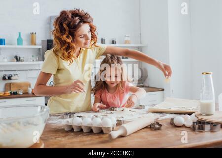 Selektiver Fokus der jungen Mutter und Tochter, die Cookies in der Nähe von Hühnereiern, Milch und Kochutensilien auf dem Küchentisch zubereiten Stockfoto