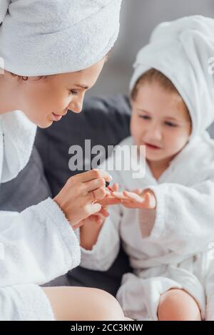 Selektiver Fokus der Frau, die Fingernägel des Kindes mit Emaille bedeckt Während Sie zusammen in weißen Bademänteln sitzen Stockfoto