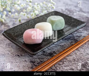 Verschiedene Geschmacksrichtungen von Mochi auf einer schwarzen rechteckigen Platte und Baby Atem Blumen Hintergrund Weichzeichnung Stockfoto