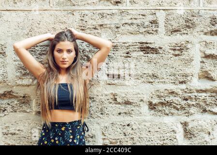 Junge blonde Mädchen in schwarzem Top und Rock mit floralem Muster, stehen mit dem Rücken an einer alten Steinwand gelehnt, ihre Arme über ihrem Kopf Blick Stockfoto