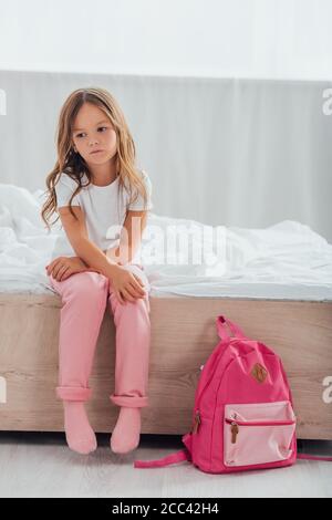 Verärgert Mädchen im Schlafanzug sitzen auf dem Bett in der Nähe Schule Rucksack Auf dem Boden Stockfoto