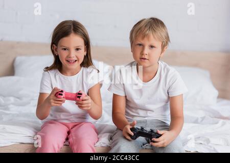 KIEW, UKRAINE - 21. JULI 2020: Aufgeregt Mädchen und konzentriert Bruder in Pyjamas spielen Videospiel im Schlafzimmer Stockfoto
