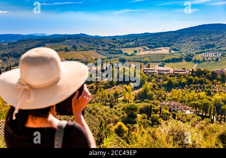 Eine Frau mit einer großen Sonnenhaube macht Fotos mit einem Handy von der schönen Landschaft von San Gimignano, Toskana. Das Bild konzentriert sich auf die ... Stockfoto