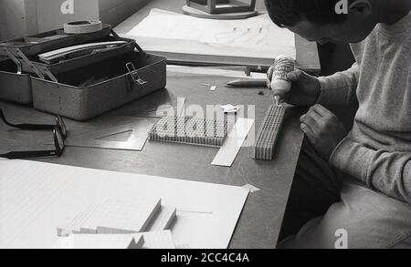 1964, Student der historischen Universitätsarchitektur, der an einem Schreibtisch sitzt und einen Krug Kleber verwendet, um ein Papier zu einem Modell zusammenzukleben, Kalifornien, USA. Stockfoto