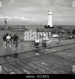 Fünfziger Jahre, historische Schüler im Teenageralter, die am Hafen, Donaghadee, Bangor, Nordirland, stehen. Der berühmte viktorianische Leuchtturm am Ende des South Pier am Eingang zum Hafen ist zu sehen. Ursprünglich 1836 erbaut, wurde der Leuchtturm im Oktober 1934 als erster in Irland auf Strom umgestellt. Ein Schild T. Simpson für Motorbootfahrten auf 'Miss Dorothy' ist am Kai zu sehen. Stockfoto