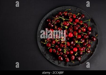 Blick von oben auf frische, nasse, dunkle Kirschen. Wassertropfen auf leuchtend dunkelroten Früchten mit Stielen. Schwarzer Steinteller auf dramatischem schwarzen Hintergrund. Stockfoto
