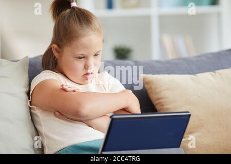 Mädchen mit Down-Syndrom sitzt auf dem Sofa mit ihren Armen Gekreuzt und etwas auf dem digitalen Tablet zu Hause anschauen Stockfoto