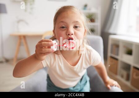 Kleines Mädchen mit Down-Syndrom Blick auf Kamera und Blasen Blasen beim Sitzen zu Hause Stockfoto