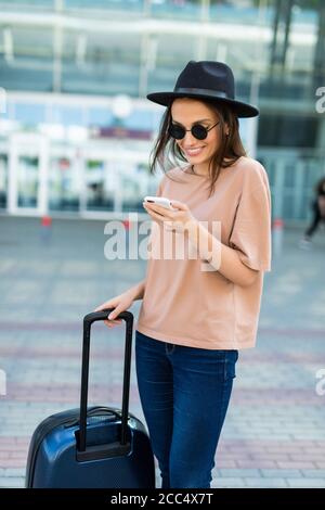 Reisende Frau mit Smartphone am Flughafen. Junge asiatische Reisende mit Handy-App im Terminal oder Bahnhof. Touristengeschäftsfrau auf dem Weg zur Arbeit Stockfoto