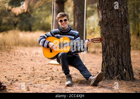 Junge schöne attraktive Mann in Pullover und schwarzen Hut Gitarre spielen im Wald sitzen auf Schaukel. Lässiger Mann, Lifestyle. Stockfoto