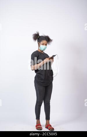 Vertikale Aufnahme einer jungen afrikanischen Dame mit einem Gesicht Maske und ihr Handy benutzen Stockfoto