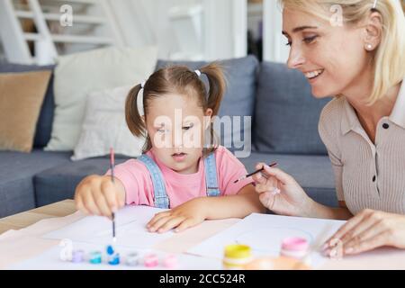 Kleines Mädchen mit Down-Syndrom lernen, an der malen Tisch mit ihrer Mutter, die ihr im Zimmer hilft Stockfoto