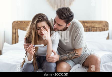 Trauriger Mann umarmt weinende Frau und schaut auf Schwangerschaftstest Im Schlafzimmer innen Stockfoto