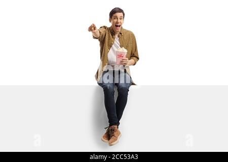 Junger Mann lachend und zeigt auf eine Tafel und isst Popcorn isoliert auf weißem Hintergrund Stockfoto