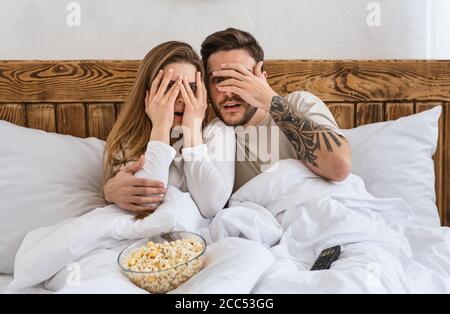 Verängstigter Ehemann und Ehefrau bedeckten die Augen mit Händen im Inneren Schlafzimmer mit Fernbedienung und Popcorn Stockfoto
