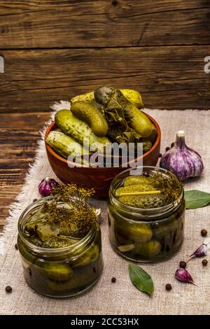 Marinierte Gurken mit Gewürzen und Kräutern. Traditionelle fermentierte Vorspeise in Gläsern und Tonschüssel. Holzbretter Hintergrund, Nahaufnahme Stockfoto