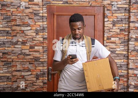 Junger afrikanischer männlicher Kurier hält das Paket, während er sein benutzt Telefon vor einem Haus Stockfoto