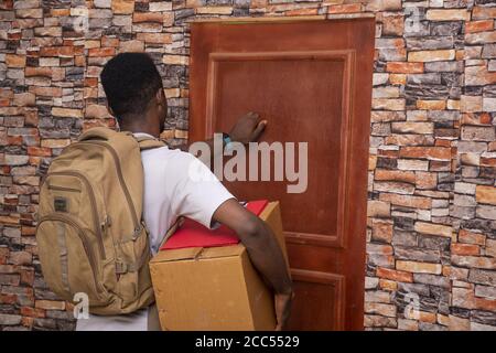 Junge afrikanische Kurier hält Paket in einer großen Box und Klopft an eine Tür Stockfoto