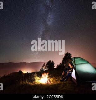 Junge romantische Paar Reisende ruhen auf Camping neben Lagerfeuer im Touristenzelt unter Nachthimmel voller Sterne und Milchstraße. Auf dem Hintergrund schöner Sternenhimmel, Berge und leuchtende Stadt Stockfoto