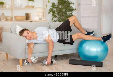 Müde Kerl mit Hanteln und Fitness-Ball liegt auf dem Sofa Mit geschlossenen Augen Stockfoto
