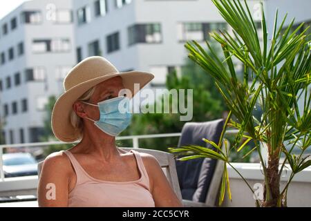 Ältere Frau mit großem Strohhut, die auf einem Balkon sitzt und eine medizinische Gesichtsmaske trägt, um sich vor Coronavirus oder Covid-19 Pandemie zu schützen. Vorderansicht des wo Stockfoto