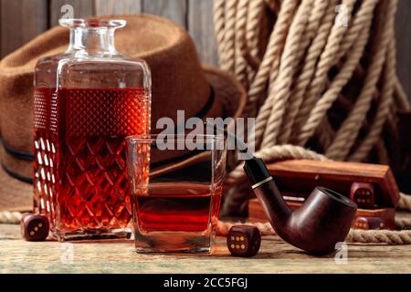 Starkes alkoholisches Getränk, Pfeife und Würfel auf einem alten Holztisch. Im Hintergrund sind ein Hanfseil und ein Männerhut. Das Konzept für die großen Jungs. Stockfoto